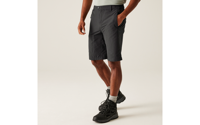 Unterkörper einer Person in dunkelgrauen knielangen Shorts, grauen Socken und schwarzen Wanderschuhen, eine Hand in der Hosentasche, heller Hintergrund