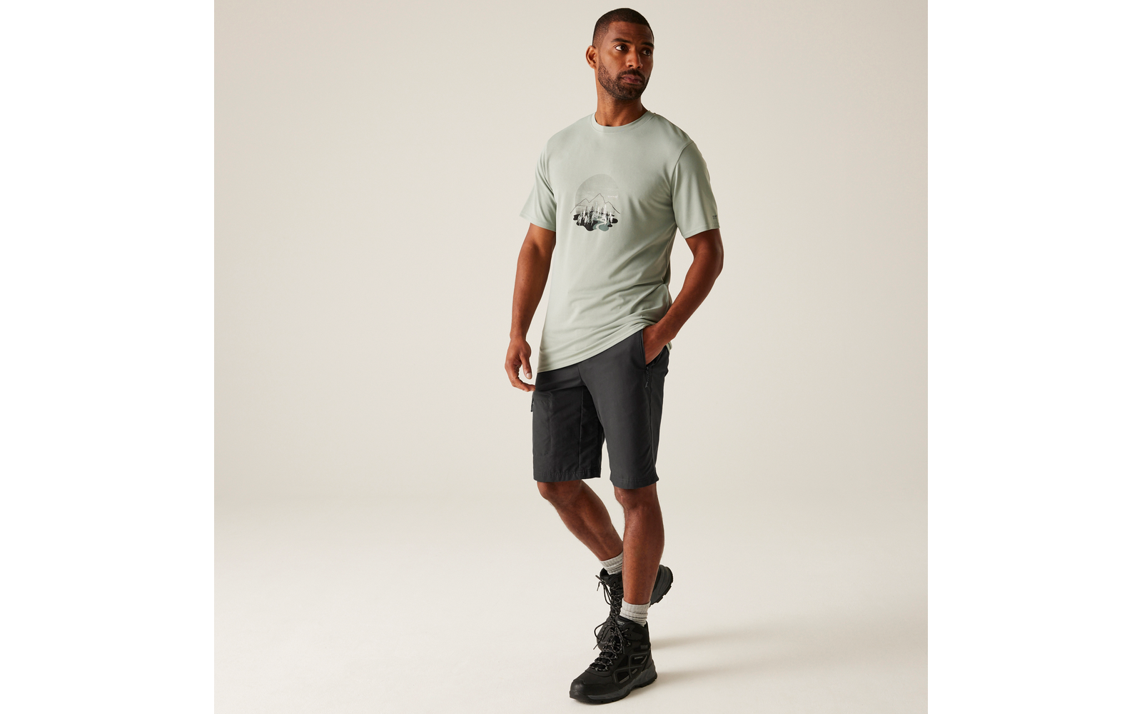 Mann steht vor hellem Hintergrund, trägt hellgrünes T‑Shirt mit rundem Print, dunkle knielange Shorts und schwarze Wanderschuhe; eine Hand in der Hosentasche