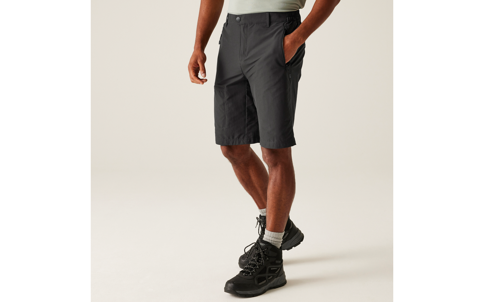 Unterkörper einer Person in dunkelgrauen knielangen Shorts, grauen Socken und schwarzen Wanderschuhen, eine Hand in der Hosentasche, heller Hintergrund