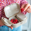 Boîte beige à compartiments ouverte; framboises dans un compartiment; main tenant une framboise au‑dessus de la boîte; torse avec haut rose à motif et jean bleu visible; visage non visible