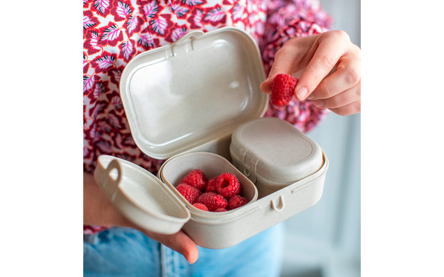 Boîte beige à compartiments ouverte; framboises dans un compartiment; main tenant une framboise au‑dessus de la boîte; torse avec haut rose à motif et jean bleu visible; visage non visible