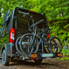 Black camper van with 'Sunlight' logo; rear bike rack holding two mountain bikes; green forest in the background; dirt ground