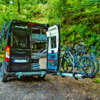Black camper van with rear doors open showing bed and storage, three bicycles on a rear rack, forested background