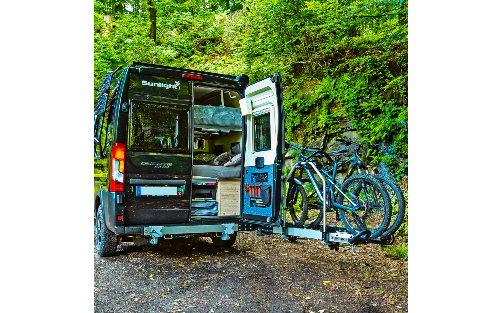 Black camper van with rear doors open showing bed and storage, three bicycles on a rear rack, forested background