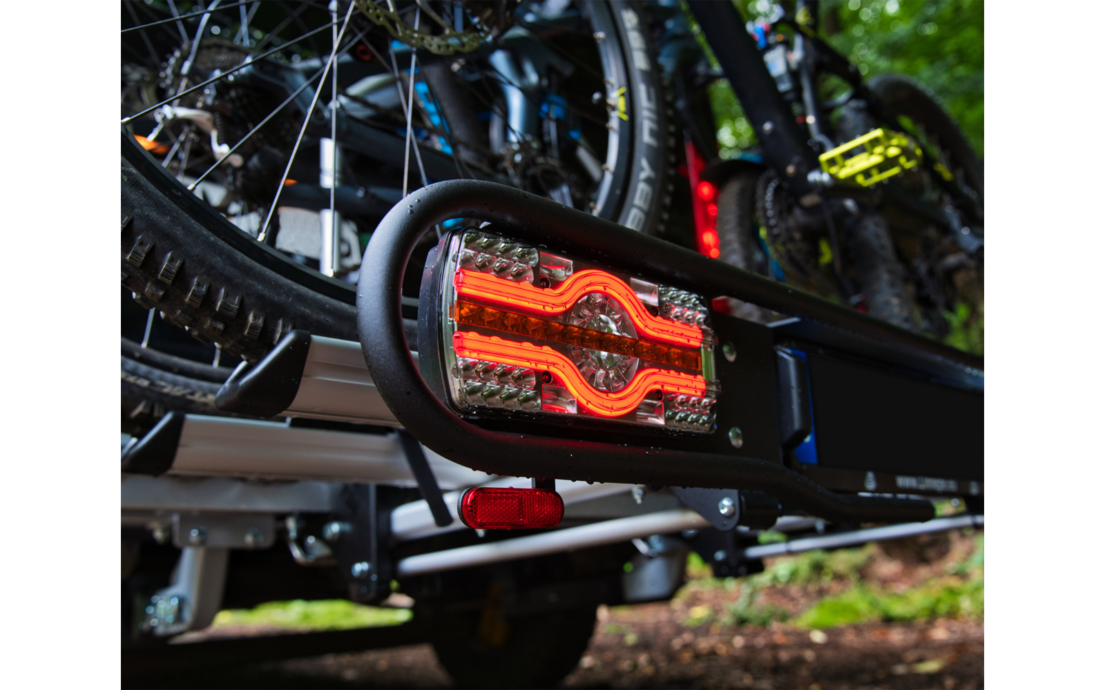 Close-up of a red illuminated bicycle rear light attached to a metal bike rack, multiple bicycles in the background