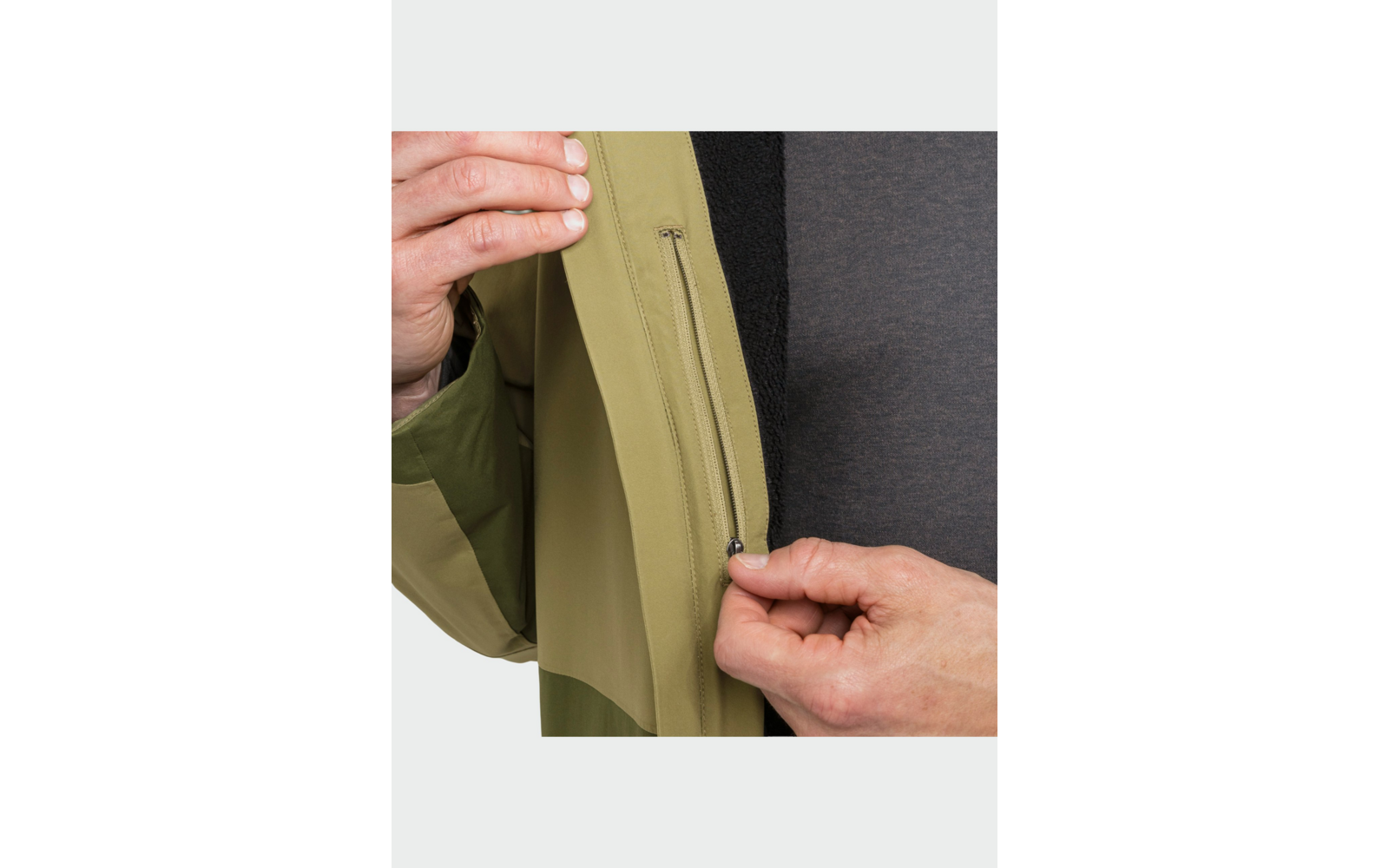 Deux mains tiennent la fermeture éclair d'une poche intérieure d'une veste vert olive; un T-shirt gris foncé est visible en dessous
