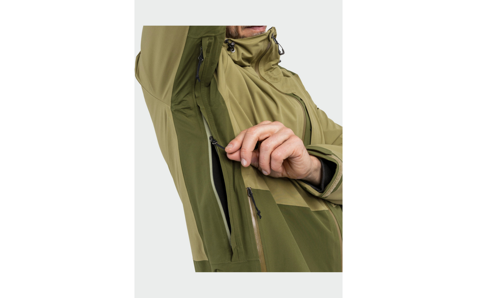 Buste d'une personne portant une veste vert bicolore, bras levé, main ouvrant une fermeture éclair sous l'aisselle ; capuche avec cordon visible