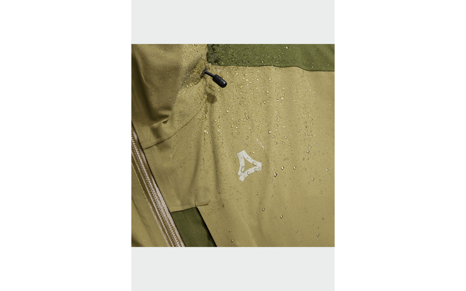 Gros plan d'une veste vert olive avec des gouttes d'eau, fermeture éclair visible, stoppeur de cordon et petit logo blanc triangulaire