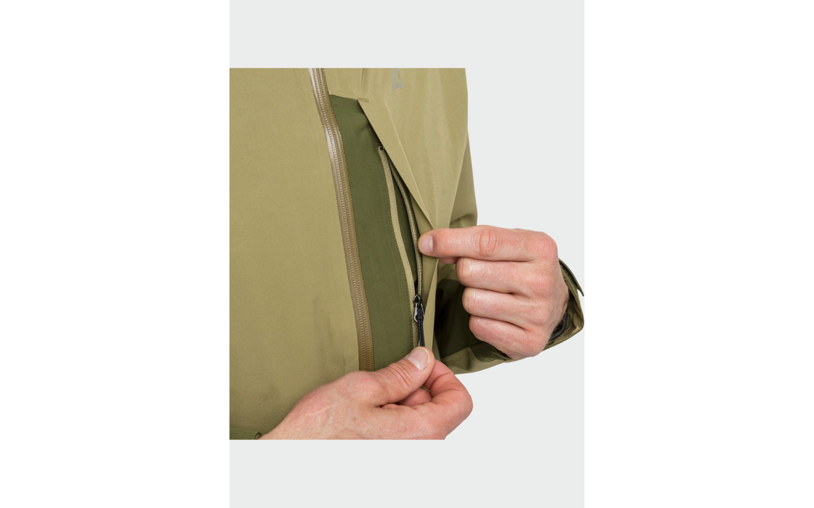 Mains ouvrant une fermeture éclair sur une veste vert olive ; fermeture intérieure et partie de la manche visibles ; fond clair