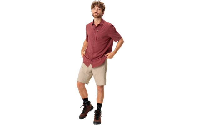 Een staande man met krullend haar en korte baard, draagt een rood gemêleerd kort mouw overhemd, beige korte broek, zwarte sokken en bruine wandelschoenen; handen in de zakken; witte achtergrond