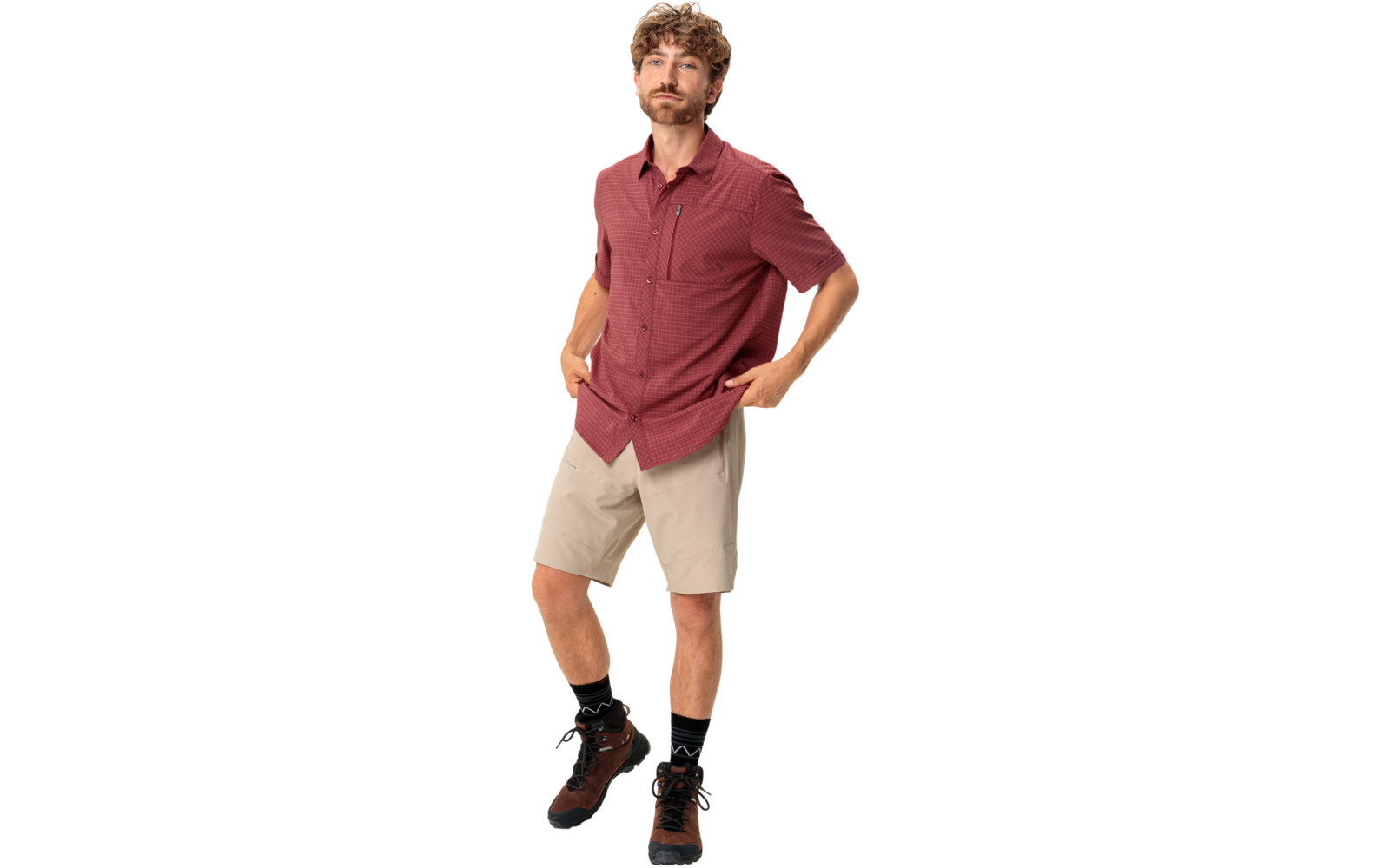 Een staande man met krullend haar en korte baard, draagt een rood gemêleerd kort mouw overhemd, beige korte broek, zwarte sokken en bruine wandelschoenen; handen in de zakken; witte achtergrond