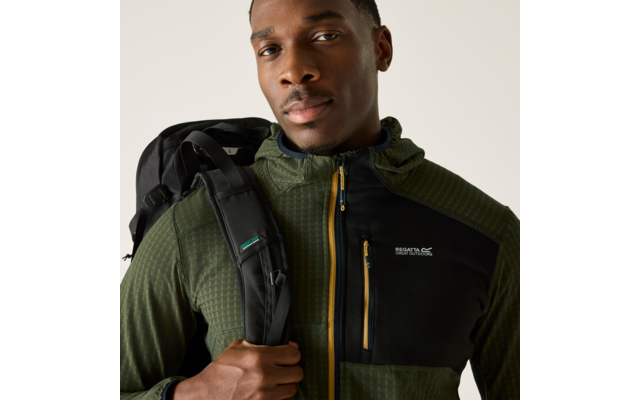 Person von der Brust aufwärts, trägt eine grüne Kapuzenjacke mit schwarzem Brustteil und gelbem Reißverschluss, hält einen Rucksackriemen, blickt zur Kamera vor hellem beige Hintergrund