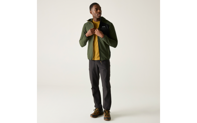 Person steht, trägt grüne Jacke, gelbes T-Shirt, dunkle Hose und braune Schuhe; Hände am Reißverschluss, Blick zur Seite