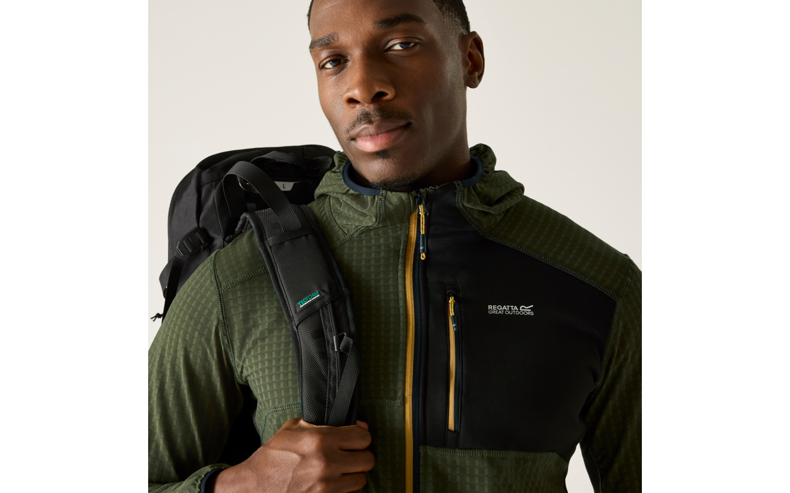 Person von der Brust aufwärts, trägt eine grüne Kapuzenjacke mit schwarzem Brustteil und gelbem Reißverschluss, hält einen Rucksackriemen, blickt zur Kamera vor hellem beige Hintergrund