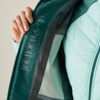Hand hält eine geöffnete dunkelgrüne Jacke; Innenseite mit weißen Piktogrammen und kleinem Text; rechts helltürkiser gesteppter Ärmel sichtbar