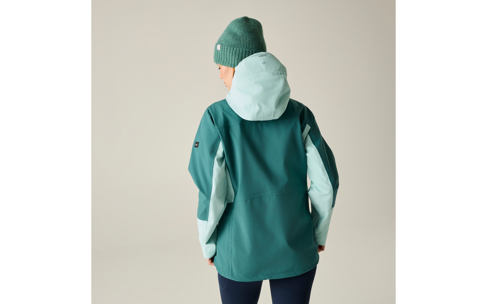 Person von hinten in grüner Jacke mit hellmintfarbener Kapuze und Ärmelfeldern, grüne Strickmütze, dunkle Hosen, heller neutraler Hintergrund