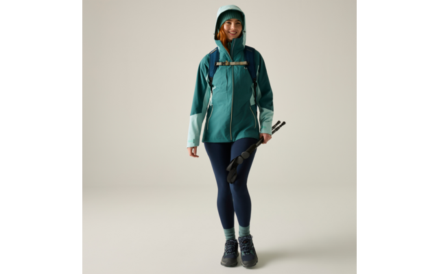 Person in türkisgrüner Jacke mit Kapuze, Rucksack, dunkelblaue Leggings, Wanderschuhe, hält zusammengeklappte Wanderstöcke, lächelt, neutraler Hintergrund
