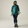 Person in türkisgrüner Jacke mit Kapuze, Rucksack, dunkelblaue Leggings, Wanderschuhe, hält zusammengeklappte Wanderstöcke, lächelt, neutraler Hintergrund