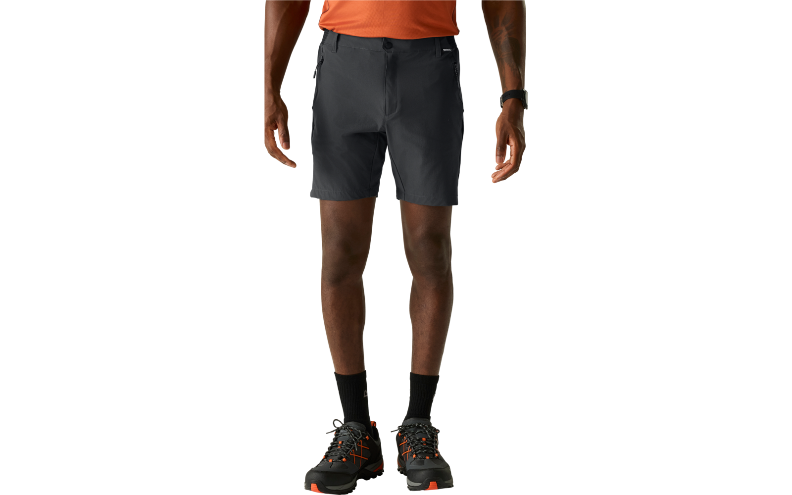 Unterkörper einer Person in dunkelgrauen Shorts, orangefarbenem Oberteil, schwarzen Socken und schwarzen Wanderschuhen mit orangefarbenen Details vor hellem Hintergrund