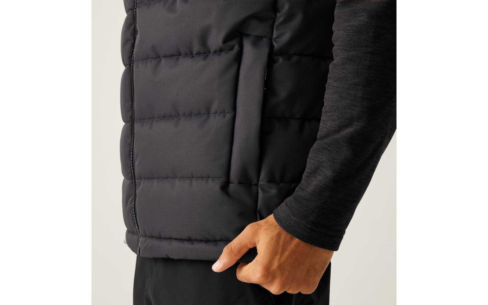 Nahaufnahme einer Hand am Saum einer schwarzen Steppweste ohne Ärmel über einem dunklen Langarmshirt und schwarzen Hosen
