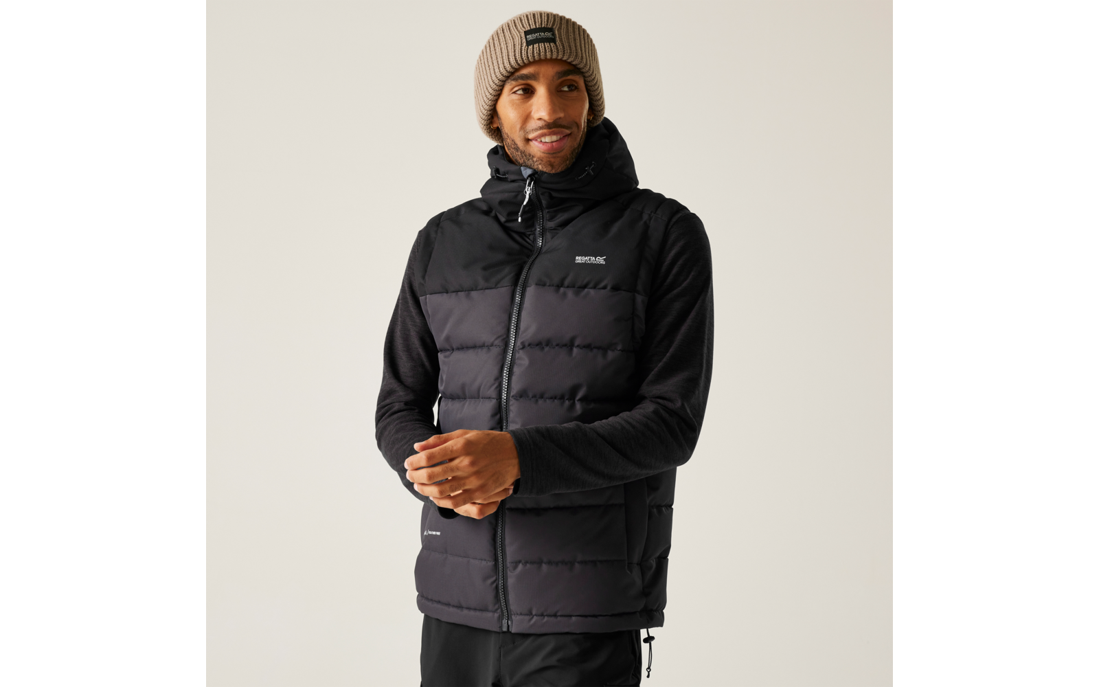 Mann trägt schwarze gesteppte Weste mit Kapuze über schwarzem Langarmshirt und beige Strickmütze, Hände vor dem Körper, heller Hintergrund