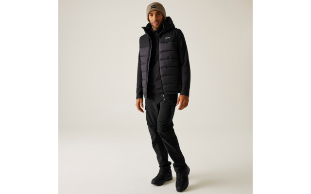 Stehende Person in schwarzer gesteppter Weste über schwarzem Hoodie, schwarzer Hose, schwarzen Schuhen und beiger Strickmütze vor hellem Hintergrund