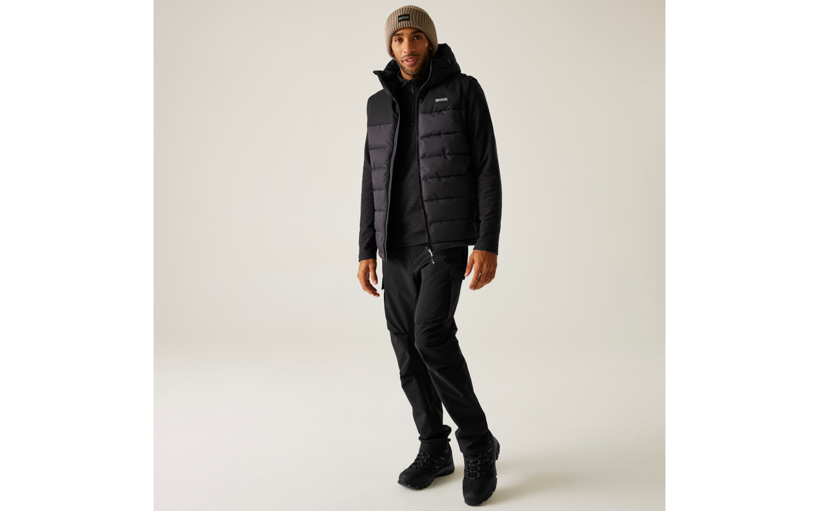 Stehende Person in schwarzer gesteppter Weste über schwarzem Hoodie, schwarzer Hose, schwarzen Schuhen und beiger Strickmütze vor hellem Hintergrund