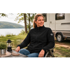 Vrouw zit op een campingstoel naast een camper aan de oever van een meer, tafel met lantaarn en onderzetters op de voorgrond, bomen op de achtergrond