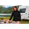 Vrouw in zwarte fleecejas en beige broek staat bij een houten tafel met fruit, glazen en servies; caravan, meer en beboste heuvels op de achtergrond