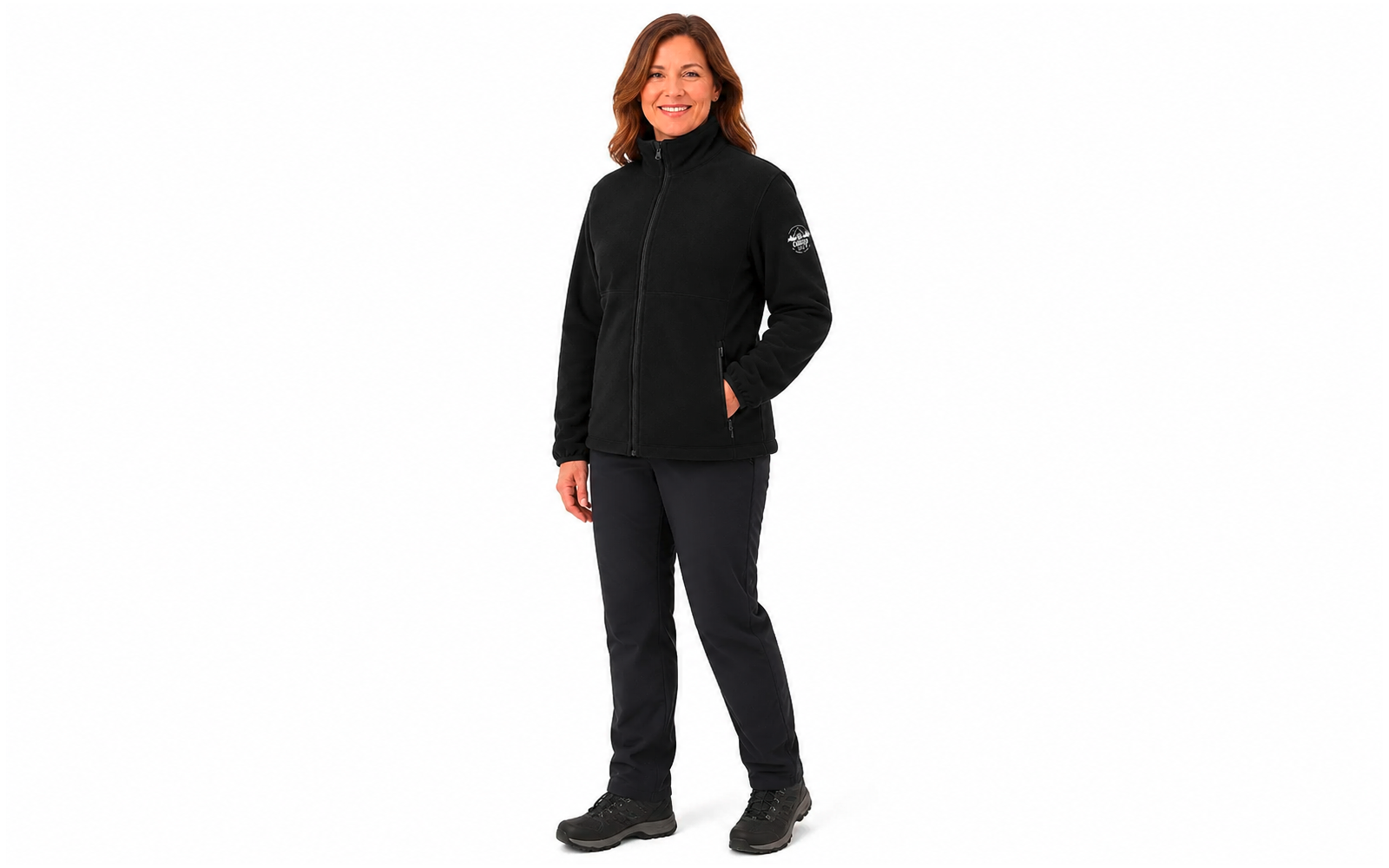 Lachende vrouw met halflang bruin haar, zwarte jas met ritssluiting, zwarte broek en zwarte schoenen, handen in de zakken, staand voor een witte achtergrond