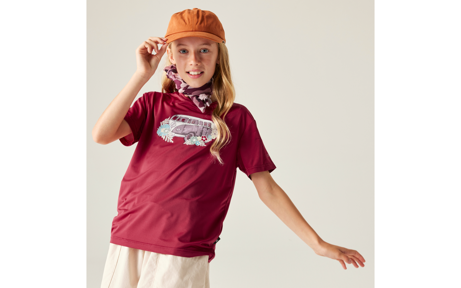 Mädchen mit orangefarbener Kappe, blondem Haar, bordeauxfarbenem T‑Shirt mit Bus‑Motiv, Halstuch und hellem Rock, eine Hand an der Kappe, andere ausgestreckt, heller Hintergrund