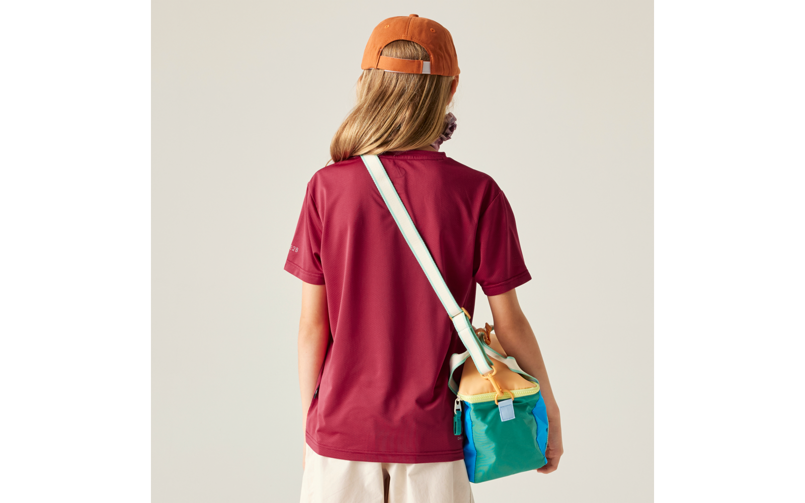 Person von hinten mit langen blonden Haaren, orangefarbener Baseballmütze rückwärts, weinrotem T‑Shirt, hellen Shorts und bunter Umhängetasche mit hellem Träger
