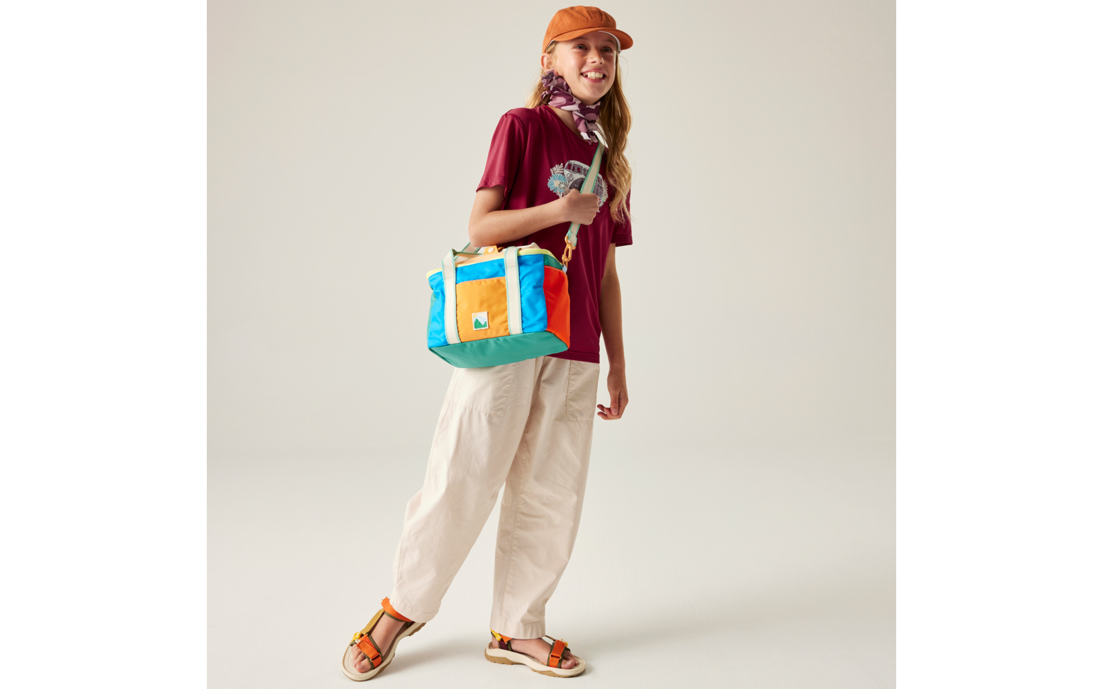 Person steht vor hellem Hintergrund, trägt orange Kappe, bordeauxrotes T‑Shirt, weite helle Hose, Sandalen, buntes rechteckiges Umhängetasche und ein Halstuch