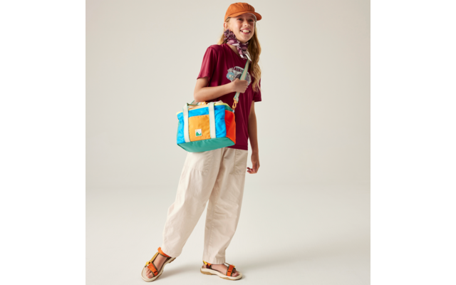 Person steht vor hellem Hintergrund, trägt orange Kappe, bordeauxrotes T‑Shirt, weite helle Hose, Sandalen, buntes rechteckiges Umhängetasche und ein Halstuch