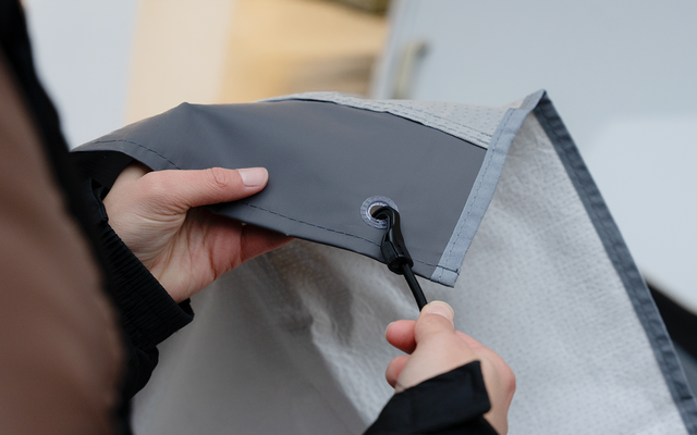 Dos manos tiran de un cordón negro con tope de plástico a través de un ojal metálico en una tela gris con interior claro