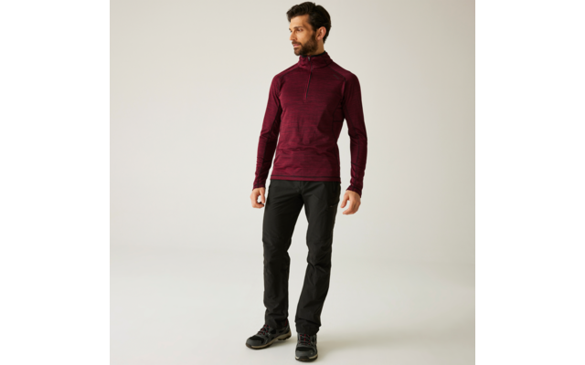 Ein Mann steht vor einem hellen Hintergrund, trägt einen roten Pullover und schwarze Hose
