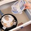 Hand holding three round blue speckled objects with white holding clips over an open drawer; inside the drawer a stainless steel pot with lid on a beige liner; drawer edges visible