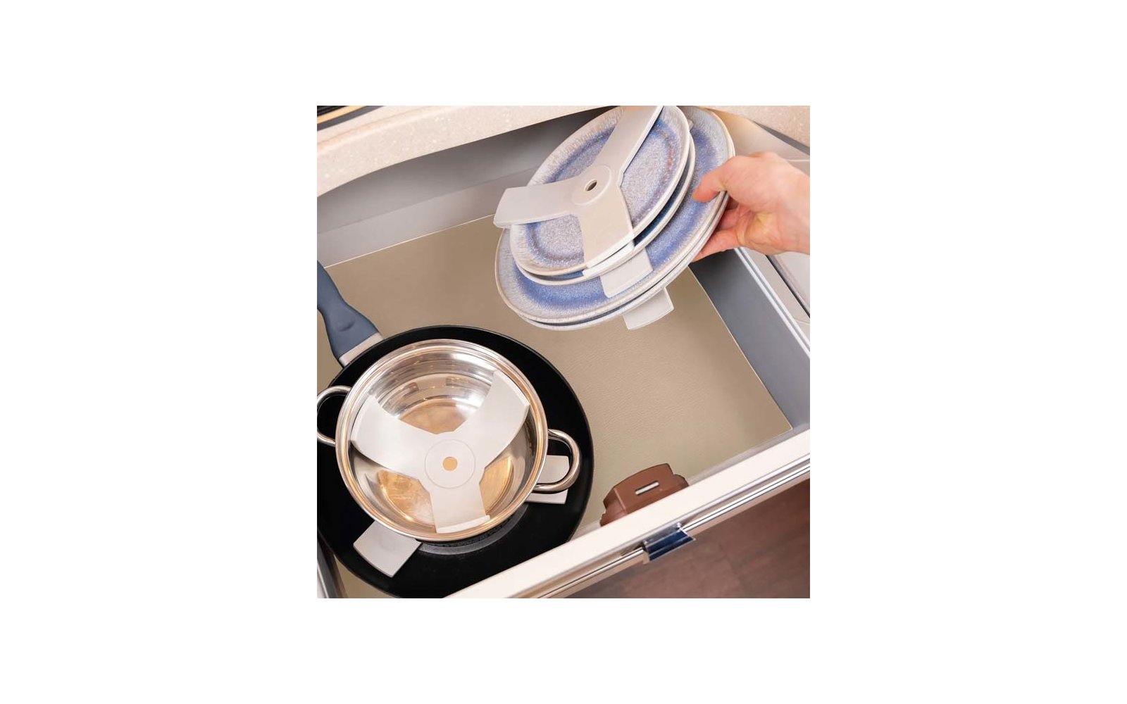 Hand holding three round blue speckled objects with white holding clips over an open drawer; inside the drawer a stainless steel pot with lid on a beige liner; drawer edges visible