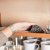 Hand with silver wristwatch touching a black packet with white pattern on a wooden shelf; below are white coffee cups and glasses