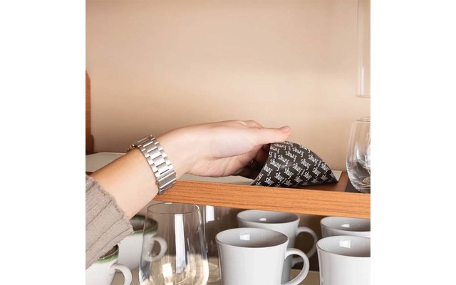 Hand with silver wristwatch touching a black packet with white pattern on a wooden shelf; below are white coffee cups and glasses