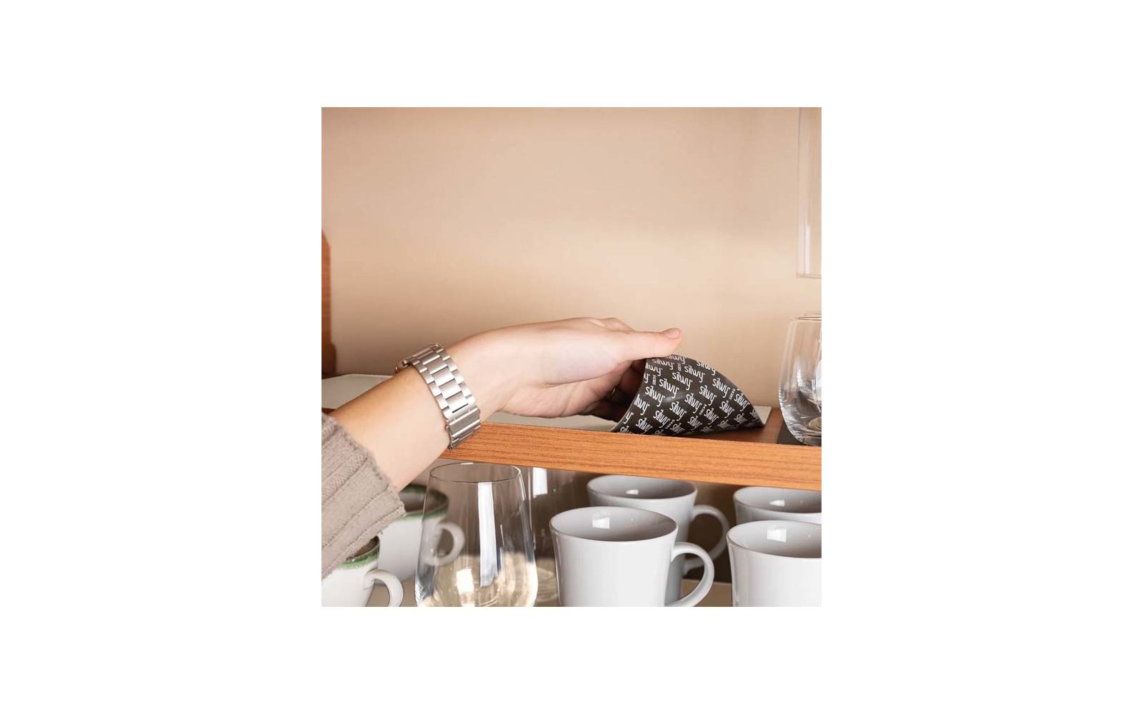 Hand with silver wristwatch touching a black packet with white pattern on a wooden shelf; below are white coffee cups and glasses
