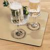 Two tall glasses with frothy pale-golden liquid on a rectangular reflective mat on a wooden table; beside them a small white vase with green leaves and small round green fruits