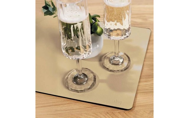 Two tall glasses with frothy pale-golden liquid on a rectangular reflective mat on a wooden table; beside them a small white vase with green leaves and small round green fruits