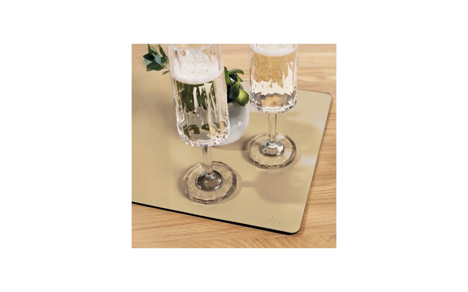 Two tall glasses with frothy pale-golden liquid on a rectangular reflective mat on a wooden table; beside them a small white vase with green leaves and small round green fruits