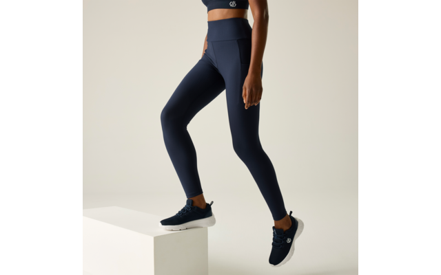 Vista parcial de una persona desde el torso hasta los pies que lleva mallas y top deportivo azul oscuro, un pie sobre un bloque blanco, zapatillas deportivas azul oscuro, fondo liso claro