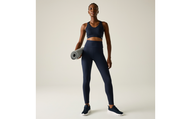 Persona de pie de frente, viste top deportivo y mallas azul marino, sostiene una esterilla de yoga enrollada en la mano derecha, zapatillas oscuras sobre fondo liso y claro