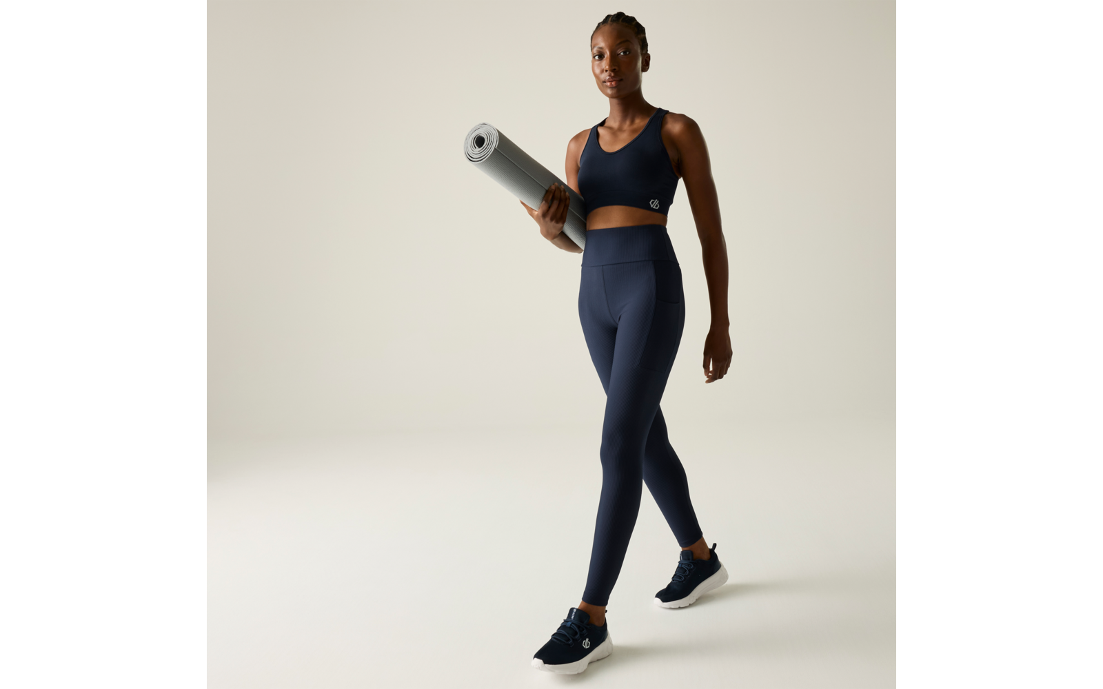 Mujer con top deportivo y mallas que lleva una esterilla de yoga enrollada, caminando frente a un fondo liso y claro