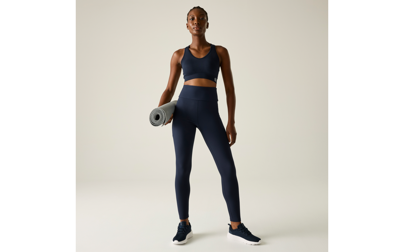 Persona de pie de frente, viste top deportivo y mallas azul marino, sostiene una esterilla de yoga enrollada en la mano derecha, zapatillas oscuras sobre fondo liso y claro