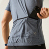 Rückenansicht einer Person in blaugrauem Sportshirt, die ein schwarzes Smartphone in eine Rückentasche steckt; sichtbarer Arm mit Armbanduhr, Logo auf dem Shirt und schwarze Sporthose