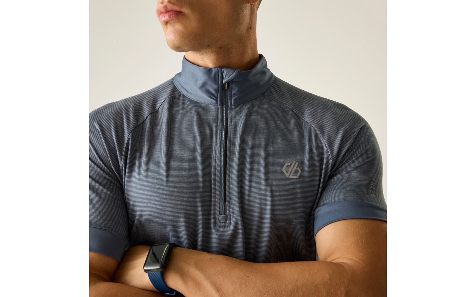 Oberkörper einer Person mit verschränkten Armen, graues kurzärmeliges Sportshirt mit kurzem Reißverschluss und kleinem Logo auf der linken Brust, Smartwatch am linken Handgelenk; nur Kinn und Unterlippe sichtbar